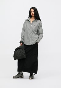 Schwarzer langer Rock mit Cargo-Taschen, kombiniert mit einem schwarz-weißen gestreiften Hemd mit Knopfleiste. Hält eine schwarze gesteppte Jacke und trägt graue Sneakers.