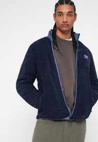 Fleece bunda v námořnické modři s texturovaným povrchem, zapínání na zip a bočními kapsami. Obsahuje modré akcenty a malé logo na levé horní části hrudi.