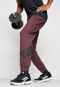 Bordeaux rode joggingbroek met een zwart logo-design met drie strepen op het linkerpootje, gecombineerd met zwarte sportschoenen en een zwarte dumbbell in de hand.