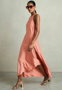 Abito maxi in satin pesca con arricciature laterali, design senza maniche e orlo asimmetrico. Abbinato a sandali con tacco a blocco dorati e occhiali da sole rossi.