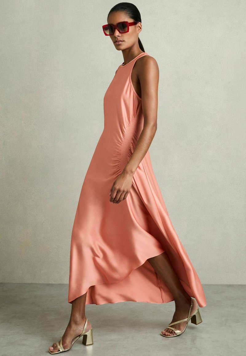 Abito maxi in satin pesca con arricciature laterali, design senza maniche e orlo asimmetrico. Abbinato a sandali con tacco a blocco dorati e occhiali da sole rossi.