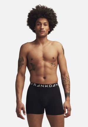 Zwarte boxer shorts met een witte gemerkte tailleband, model met tatouages op beide armen, staat met een neutrale uitdrukking tegen een effen achtergrond.