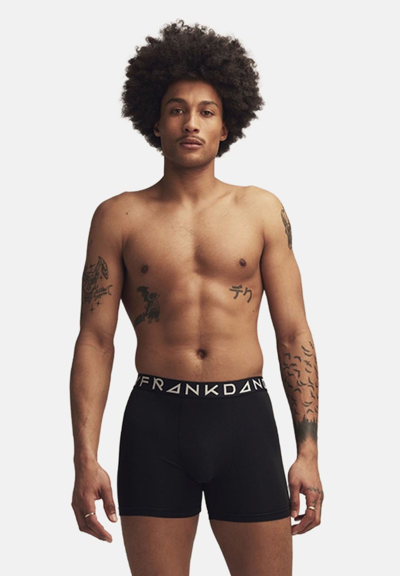 Svarta boxerkalsonger med en vit märkesmidja, modellen har tatueringar på båda armarna och står med ett neutralt uttryck mot en enfärgad bakgrund.
