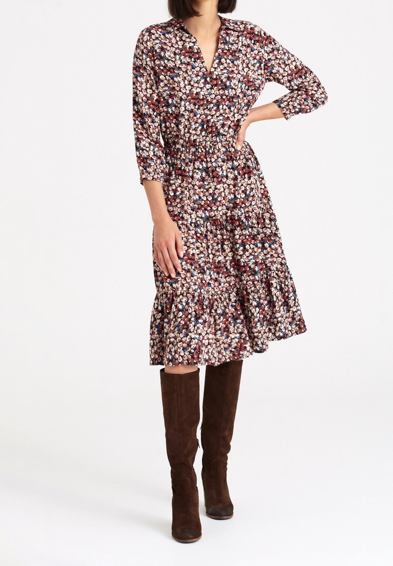 Femme portant une robe midi à manches longues fleurie, avec une taille froncée et des bottes en daim marron montant jusqu'aux genoux, debout sur un fond blanc.
