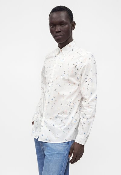 Homme portant une chemise blanche boutonnée avec de petits motifs abstraits multicolores et un jean bleu clair, debout devant un fond uni.