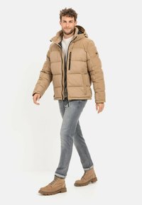 Beige Pufferjacke mit Kapuze, Reißverschluss und seitlichen Taschen. Getragen über einem grauen Pullover und kombiniert mit hellblauen Jeans und beigen Stiefeln.