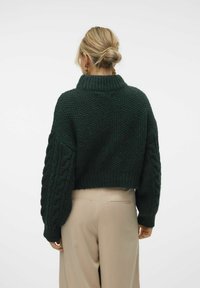 Groene gebreide cropped trui met hoge kraag en kabelbreipatronen op de mouwen, gecombineerd met beige high-waisted broek.