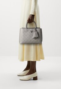 Borsa grigia con texture e motivi in rilievo, con doppio manico, un'etichetta staccabile e una forma rettangolare strutturata. Indossata con un vestito giallo.