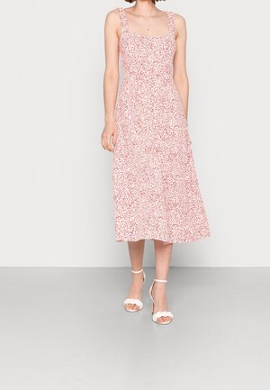 Vrouw draagt een mouwloze, roze midi-jurk met rode stippen en witte sandalen met hoge hakken, staand tegen een effen achtergrond.