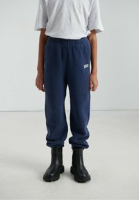 Tumši zili sporta bikses ar elastīgu jostu un aprocētām potītēm, ar mazu baltu apdruku uz kreisā augšstilba. Valdātas kopā ar melniem potīšu zābakiem.