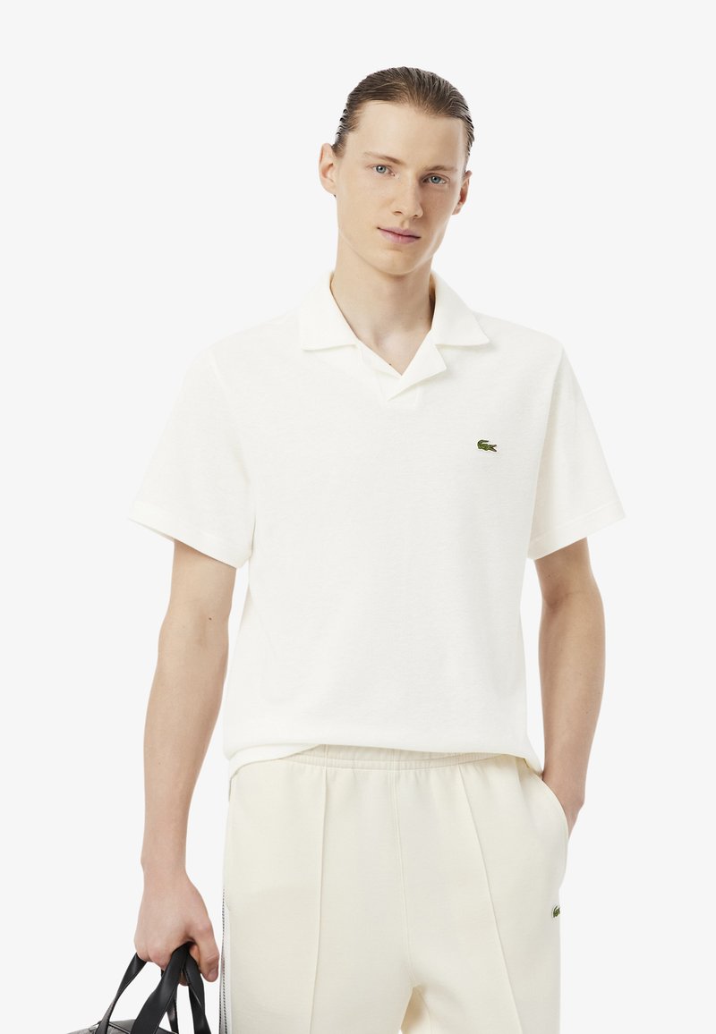 Joven con el cabello peinado hacia atrás, lleva una camiseta polo blanca y pantalones color crema, sosteniendo una bolsa negra con la mano izquierda.