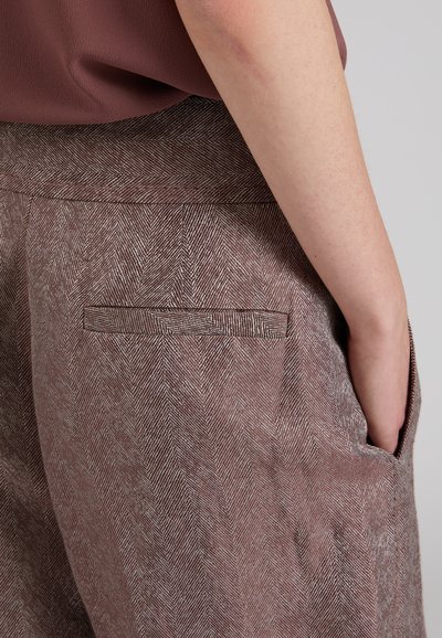 Primo piano di una persona che indossa pantaloni marroni con motivo a spina di pesce, con una tasca posteriore a filetto e la mano destra nella tasca laterale.