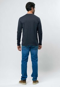 Homme debout, de dos, portant une chemise noire à manches longues, un jean bleu et des chaussures marron, sur un fond clair uni.