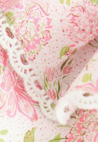 Detailný záber na mäkkú bielu látku s ružovými a zelenými kvetinovými vzormi a bielou háčkovanou čipkou na okraji.