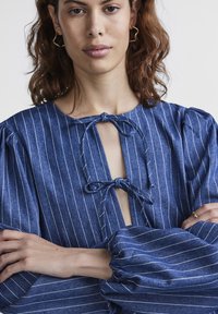 Μπλε ριγέ τζιν μπλούζα με λαιμόκοψη τύπου κλειδώματος, δεμένη με το ίδιο ύφασμα, και φουσκωτά μανίκια με ελαστικούς σφιγκτήρες για εφαρμοστή υφή.