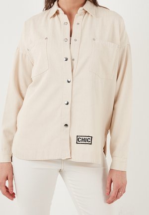 Beżowa koszula na długi rękaw zapinana na guziki z dwoma kieszeniami na piersi i naszywką "CHIC", noszona z białymi spodniami na osobie.
