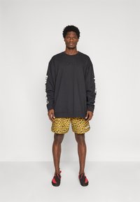 Juodas didelio dydžio džemperis su logotipu, derinamas su geltonos spalvos leopardo rašto šortais. Modelis dėvi raudonus ir juodus batus, stovi ant paprasto fono.