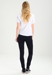 Camiseta de algodón blanca con mangas cortas, combinada con jeans ajustados de color azul oscuro y zapatillas Vans negras, vista desde la parte de atrás.