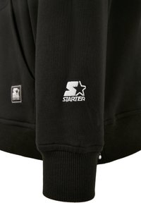 Manche de sweat-shirt noire avec bord-côtes et logo blanc Starter brodé près du poignet.