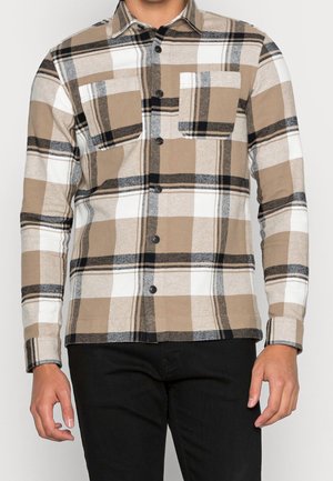 Mann trägt ein beige-weiß-schwarz kariertes Hemd mit langen Ärmeln und Knöpfen, das zwei Brusttaschen hat, sowie eine schwarze Hose.