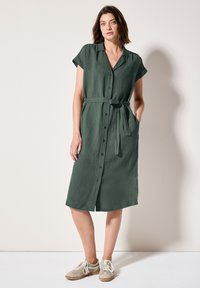 Abito verde con maniche corte e bottoni, dotato di una vita con cintura, realizzato in tessuto leggero. Presenta un colletto e tasche laterali. Abbinato a sneakers.
