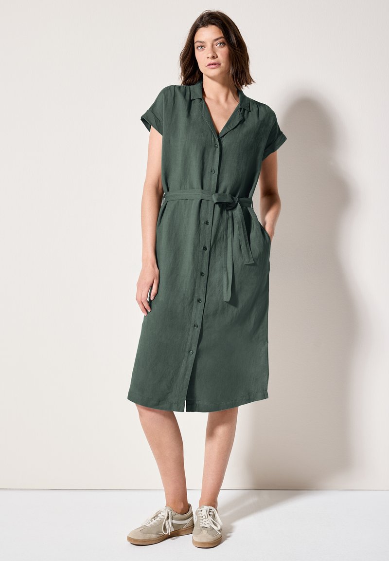 Abito verde con maniche corte e bottoni, dotato di una vita con cintura, realizzato in tessuto leggero. Presenta un colletto e tasche laterali. Abbinato a sneakers.