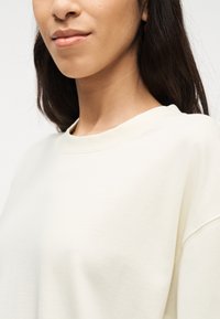 Γυναίκα με μεσαία επιδερμίδα και σκούρα μαλλιά, που φοράει απλό λευκό μπλουζάκι με στρογγυλή λαιμόκοψη, το οποίο έχει κοπεί στο ύψος του πηγουνιού και των ώμων.