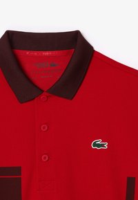 Röd polo t-shirt med en mörkbrown krage, med två knappar, en grön krokodil-logotyp och detaljer i texturerad tyg.