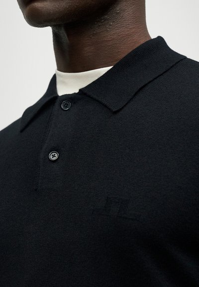 Close-up de uma pessoa a usar uma camisa polo preta com botões, por cima de uma t-shirt branca, mostrando o colarinho e a área superior do peito.