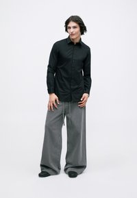 Cămașă neagră cu nasturi, cu mâneci lungi și o textură netedă, asortată cu pantaloni largi din denim gri, cu șnur. Pantofi negri.