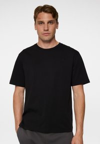 Mustapaitainen lyhythihainen T-paita puuvillamateriaalista, jossa on klassinen pyöreä kaula-aukko ja hillitty logo-yksityiskohta rinnassa.