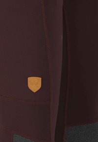 Leggings de sport bordeaux avec un panneau texturé noir, coutures surpiquées et un patch en cuir beige portant la marque "WHISTLER". Tissu lisse et extensible.