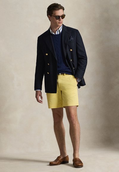 Námornícky dvojradový sako, pod ním textúrovaný sveter, pruhovaná košeľa, žlté šortky, hnedé mokasíny a tmavé slnečné okuliare.