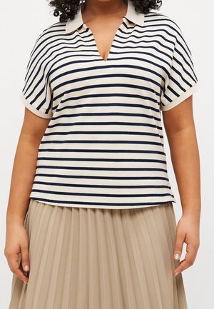 Persoon draagt een beige shirt met korte mouwen en donkerblauwe horizontale strepen en een beige geplooide rok, staand met ontspannen armen naast het lichaam.