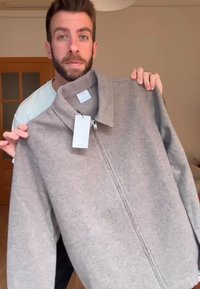 Svetlosivá bunda na zips vyrobená z mäkkého fleecového materiálu, s vysokým stojačikom a štítkom pripevneným pri zipsu.