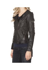 Femme portant une veste en cuir noire ajustée avec des détails zippés sur une chemise marine et un jean foncé, debout, les bras détendus le long du corps.