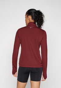 Bordo dug mad dugi rukav, sportskog kroja s visokim izrezom, rupama za palce i Nike logotipom na leđima. Uparen s crnim šorcima. Glatka, rastezljiva tkanina.