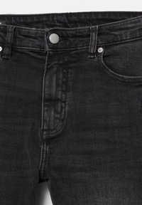 Crne traperice od trapera s prednjim gumbom, petljama za remen i vidljivim šavovima na džepovima i pojasu.