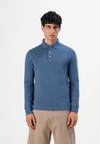 Mann in einem blauen Langarm-Poloshirt mit drei Knöpfen und beigen gestreiften Hosen, der vor einem schlichten weißen Hintergrund steht.