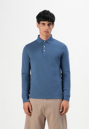 Mand iført en blå langærmet polo skjorte med tre knapper og beige stribede bukser, stående mod en helt hvid baggrund.