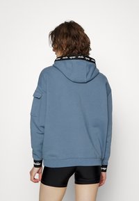 Sweat à capuche bleu avec un col noir logoé, design oversized et une poche sur la manche gauche. Comprend des poignets et une taille côtelés.