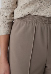 Beige gebreide trui gecombineerd met lichtbruine textuurbroek met een elastische tailleband en subtiele plooien. Close-up toont de details van de stof.