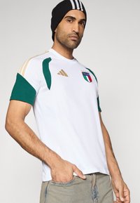 Hvid sportstrøje med grønne og gyldne detaljer, med det italienske landsholdets logo og trekantet design på ærmerne.