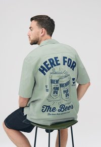 Light green short-sleeve shirt featuring navy graphic design on the back with beer cans and text "HERE FOR THE BEER." Casual fit, smooth texture.