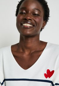 Pull en V blanc avec des rayures navy et un cœur brodé rouge sur le côté gauche. Texture tricot douce, coupe décontractée.