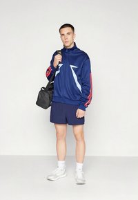 Mörkblå zip-up träningsjacka med röda ränder, ljusblå detaljer och matchande mörkblå shorts. Modellen håller en liten svart väska och bär vita sneakers.