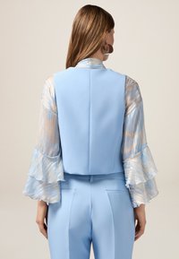 Gilet azzurro su misura abbinato a maniche ampie e trasparenti con motivi a spirale in tonalità pastello. Tessuto liscio con minimo hardware.