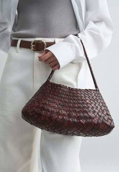 Sac à main en cuir tressé marron avec une forme courbée, une courte anse et une surface texturée, associé à un haut gris et un pantalon blanc.