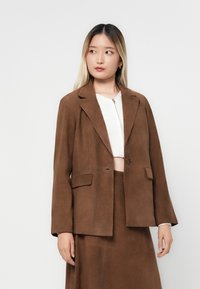 Blazer en daim marron avec un col cranté, fermeture à un bouton, et deux poches avant. Fini texturé et coupe légèrement surdimensionnée.