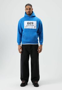 Sweat-shirt à capuche bleu avec un grand écusson graphique blanc portant l'inscription "009", associé à un pantalon noir ample et des chaussures noires à lacets.